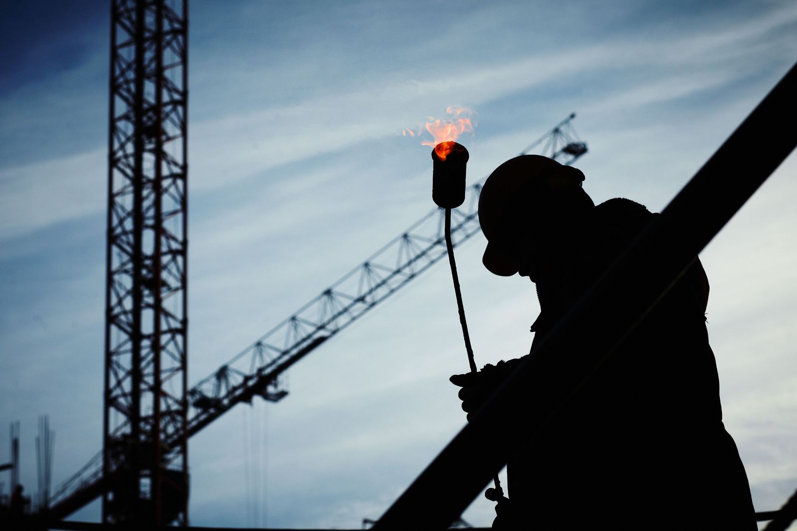 Silhouet van een arbeider met een snijbrander op een werf, met bouwkranen op de achtergrond